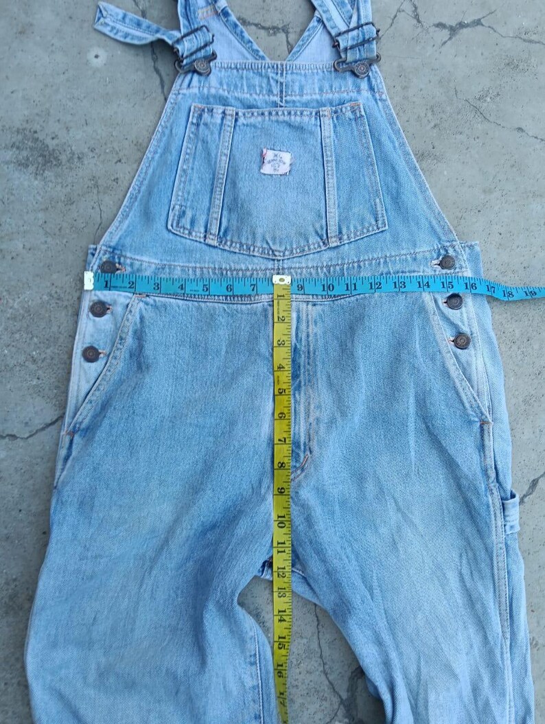 Op de afbeelding: Lichtblauwe denim overall met verstelbare bandjes, meerdere zakken en knoopsluitingen. Een meetlint geeft aan dat de taille ongeveer 35,5 cm breed is. De overall heeft een kleine patch op de borstzak.