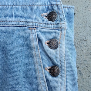 Op de afbeelding: Close-up van een lichtblauwe denim overall. De overall heeft drie donkere metalen knopen aan de zijkant en zichtbare stiksels. De denim heeft een vervaagd uiterlijk, wat wijst op een vintage of versleten look. Het algehele ontwerp is eenvoudig en klassiek.