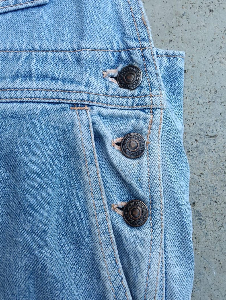 Op de afbeelding: Close-up van een lichtblauwe denim overall. De overall heeft drie donkere metalen knopen aan de zijkant en zichtbare stiksels. De denim heeft een vervaagd uiterlijk, wat wijst op een vintage of versleten look. Het algehele ontwerp is eenvoudig en klassiek.