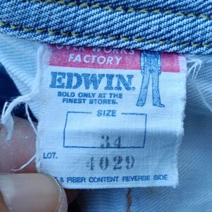 Op de afbeelding: Close-up van een wit label van Edwin jeans. Het label toont "OVER WORKS FACTORY" en "EDWIN" in vette letters. De maat is aangegeven als 34, en het lotnummer is 4029. Het label is bevestigd aan blauwe denim stof.