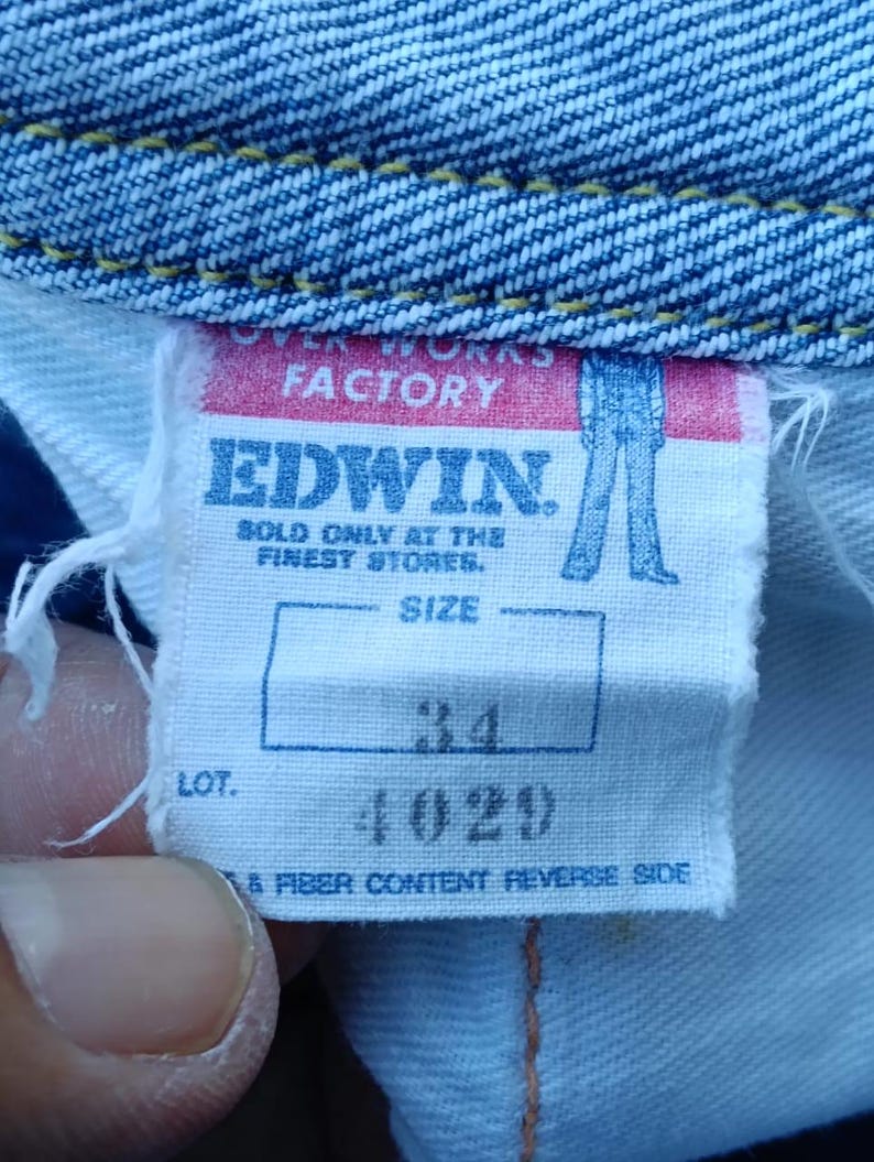 Op de afbeelding: Close-up van een wit label van Edwin jeans. Het label toont "OVER WORKS FACTORY" en "EDWIN" in vette letters. De maat is aangegeven als 34, en het lotnummer is 4029. Het label is bevestigd aan blauwe denim stof.
