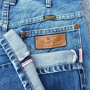 Pu&ograve; includere: Primo piano di jeans di denim blu con il logo Wrangler sulla tasca posteriore e sulla cintura. I jeans hanno un design classico con un risvolto piegato, che mostra strisce rosse e bianche all'interno.