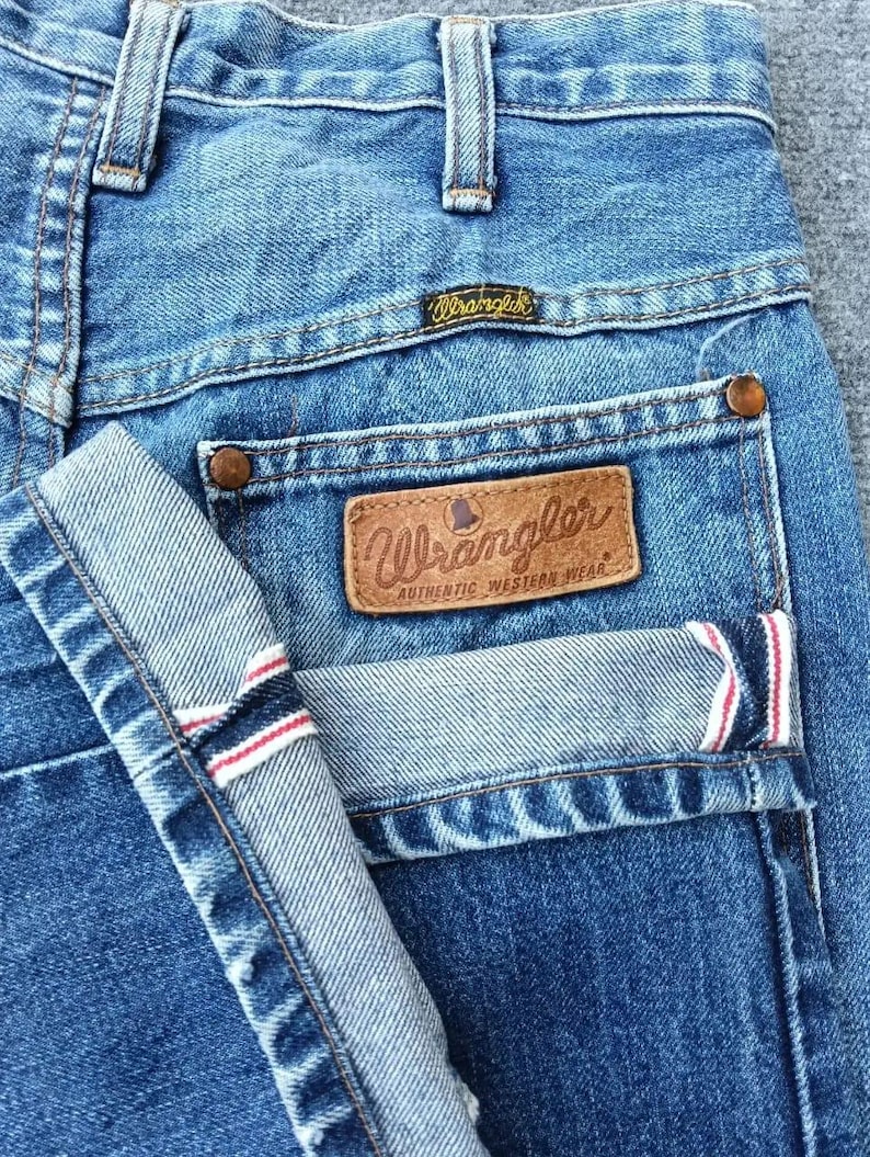 Pu&ograve; includere: Primo piano di jeans di denim blu con il logo Wrangler sulla tasca posteriore e sulla cintura. I jeans hanno un design classico con un risvolto piegato, che mostra strisce rosse e bianche all'interno.
