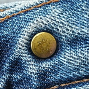 Pu&ograve; includere: Primo piano di un bottone in ottone su tessuto denim blu. Il bottone &egrave; leggermente ossidato e presenta testo in rilievo. Il denim ha cuciture visibili con filo arancione-marrone a contrasto, tipiche dei jeans.