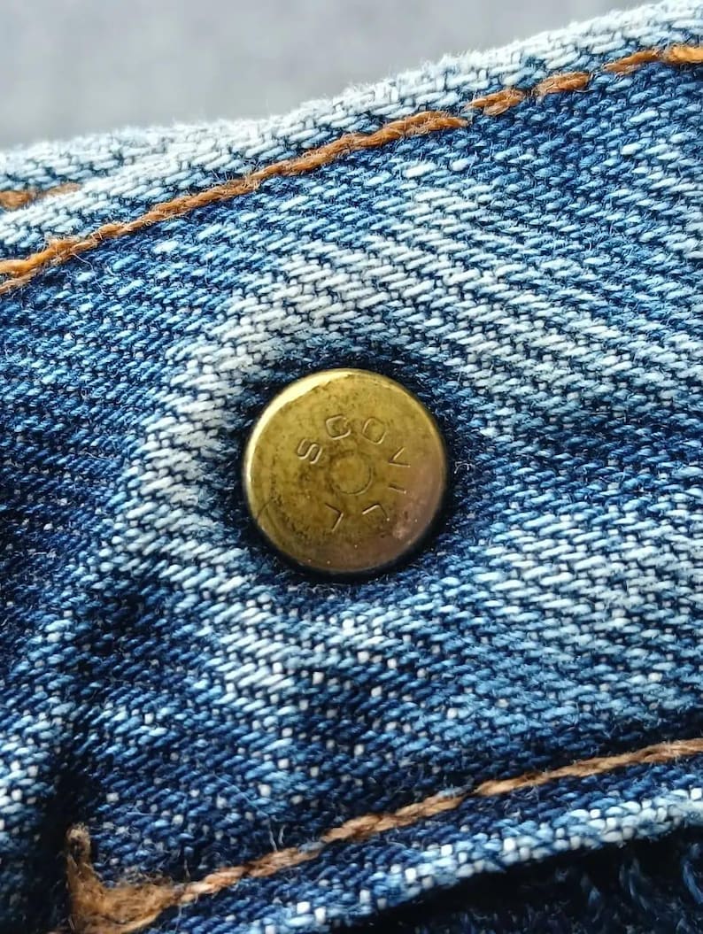 Pu&ograve; includere: Primo piano di un bottone in ottone su tessuto denim blu. Il bottone &egrave; leggermente ossidato e presenta testo in rilievo. Il denim ha cuciture visibili con filo arancione-marrone a contrasto, tipiche dei jeans.