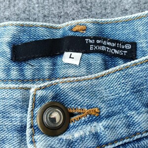 Puede incluir: Primer plano de unos vaqueros de mezclilla azul claro. Los vaqueros tienen cierre de bot&oacute;n y una etiqueta negra con el texto "The original ttu EXHIBITIONIST" y una talla "L". Las costuras son de color marr&oacute;n claro en contraste.