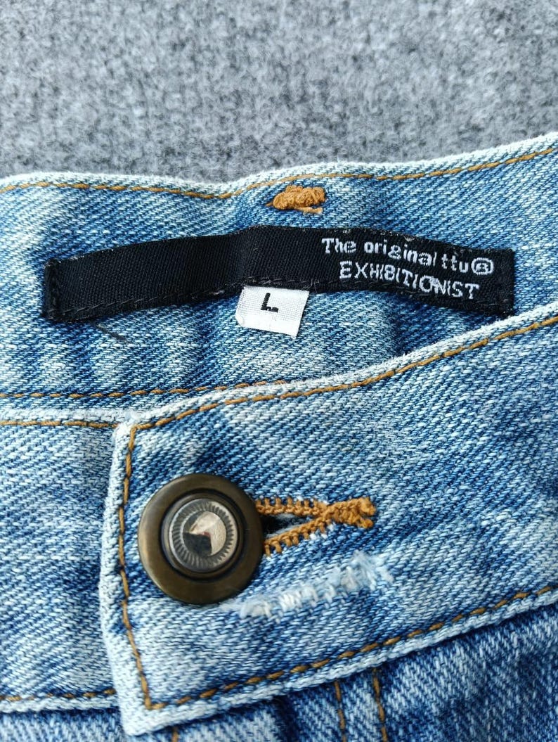 Puede incluir: Primer plano de unos vaqueros de mezclilla azul claro. Los vaqueros tienen cierre de bot&oacute;n y una etiqueta negra con el texto "The original ttu EXHIBITIONIST" y una talla "L". Las costuras son de color marr&oacute;n claro en contraste.