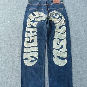 Puede incluir: Pantalones vaqueros de mezclilla azul oscuro con un estampado gráfico grande, desgastado y de color claro que se extiende por ambas piernas traseras. El diseño incluye letras estilizadas que dicen MIGHTY en la pierna izquierda y MIGHTY en la derecha. Un parche de cuero marrón es visible en la cintura trasera.