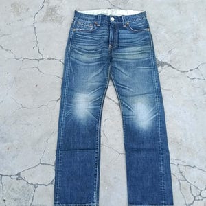 May include: A pair of blue denim jeans featuring a faded wash, whiskering patterns at the hips, and lightened areas at the knees. The jeans have a standard button fly closure, five-pocket construction, and a straight leg cut with visible stitching.