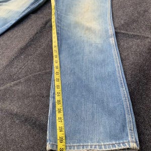 Puede incluir: Vaqueros de mezclilla azul claro con pernera acampanada, medidos con una cinta m&eacute;trica amarilla. Los vaqueros miden aproximadamente 79 cm de largo.