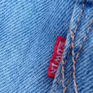 Op de afbeelding: Close-up van lichtblauwe denimstof met zichtbare textuur en stiksels. Een klein rood label met de merknaam "Edwin" is in de naad genaaid. De denim heeft een klassieke, versleten uitstraling.