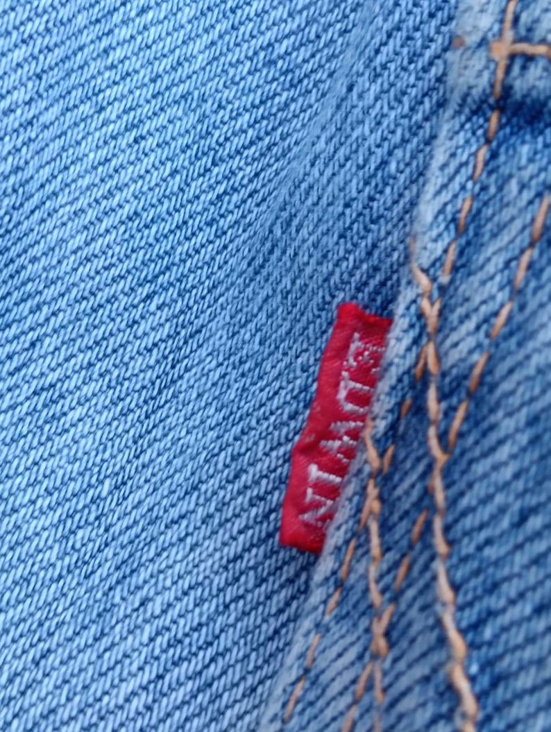 Op de afbeelding: Close-up van lichtblauwe denimstof met zichtbare textuur en stiksels. Een klein rood label met de merknaam "Edwin" is in de naad genaaid. De denim heeft een klassieke, versleten uitstraling.