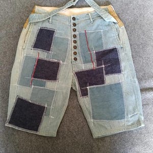 Op de afbeelding: Lichtblauwe denim shorts met een patchworkontwerp van donkerblauwe rechthoekige stoffen panelen, zichtbare witte en rode stiksels, een knoopsluiting met zeven donkere knopen en een bijpassende denim riemlusband aan de tailleband.