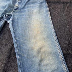 Puede incluir: Vaqueros de mezclilla azul claro con un dise&ntilde;o cl&aacute;sico de cinco bolsillos. Los vaqueros muestran signos de desgaste con &aacute;reas descoloridas y una sutil decoloraci&oacute;n. El corte recto y la construcci&oacute;n robusta sugieren un estilo vintage o de trabajo.