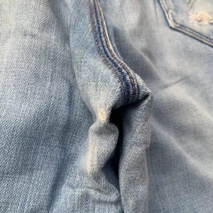 Puede incluir: Primer plano de unos vaqueros de mezclilla azul claro, que muestran la textura de la tela y los detalles de las costuras. La imagen destaca las costuras y la zona del bolsillo, con un enfoque en el aspecto desgastado y descolorido de la mezclilla.