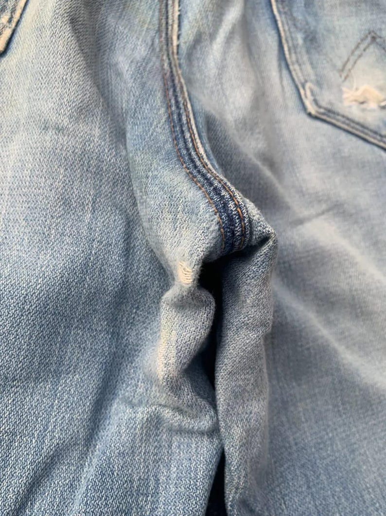 Puede incluir: Primer plano de unos vaqueros de mezclilla azul claro, que muestran la textura de la tela y los detalles de las costuras. La imagen destaca las costuras y la zona del bolsillo, con un enfoque en el aspecto desgastado y descolorido de la mezclilla.