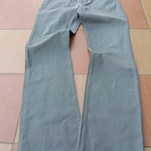 Può includere: Un paio di jeans verde oliva. I jeans hanno un classico design a cinque tasche, una chiusura con bottone e cerniera e una gamba svasata. Il tessuto sembra essere un denim di peso medio. Ci sono alcune piccole macchie bianche sulla gamba destra.