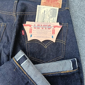 Könnte beinhalten: Dunkelblaue Jeans aus Indigo-Denim mit einem hellbraunen Lederaufnäher am Bund mit der Aufschrift Levi Strauss & Co. 501 XX W31 L36. Eine rote Lasche befindet sich an der Gesäßtasche, die zwei Papieranhänger mit dem Text The Rivet's Still There enthält.