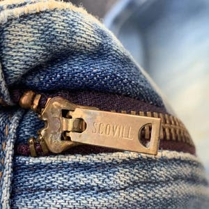 Puede incluir: Primer plano de una prenda vaquera con una cremallera de color lat&oacute;n. El tirador de la cremallera est&aacute; marcado con el nombre de la marca "SCOVILL". El tejido vaquero es de color azul descolorido, con desgaste visible.