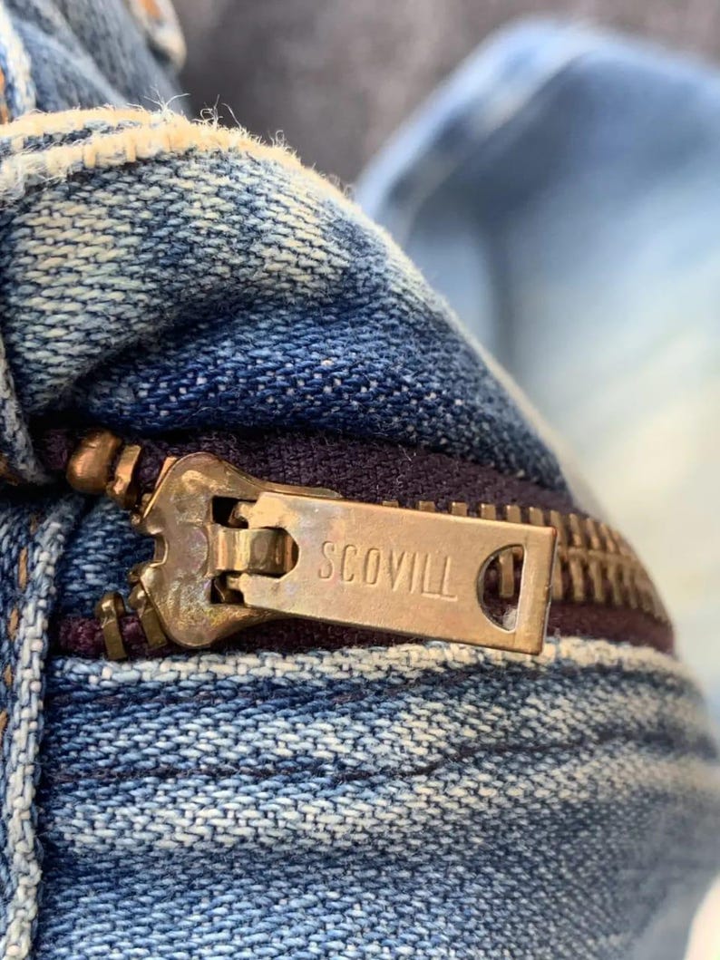 Puede incluir: Primer plano de una prenda vaquera con una cremallera de color lat&oacute;n. El tirador de la cremallera est&aacute; marcado con el nombre de la marca "SCOVILL". El tejido vaquero es de color azul descolorido, con desgaste visible.