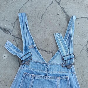Op de afbeelding: Lichtblauwe denim overall met verstelbare bandjes en metalen sluitingen. Op de borstzak zit een klein, geborduurd label. De overall ligt plat, wat het klassieke ontwerp benadrukt.