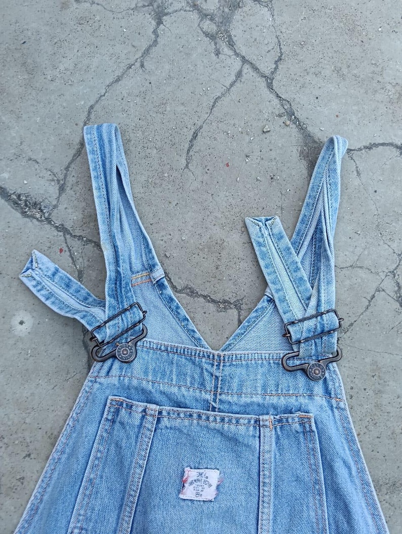 Op de afbeelding: Lichtblauwe denim overall met verstelbare bandjes en metalen sluitingen. Op de borstzak zit een klein, geborduurd label. De overall ligt plat, wat het klassieke ontwerp benadrukt.