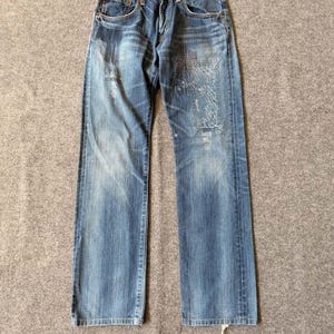 Könnte beinhalten: Blaue Jeans aus Denim mit verwaschener Optik, Knopfleiste und sichtbaren, geflickten Stellen mit aufwendigen Nähten auf den Vorderseiten. Die Jeans hat ein klassisches Fünf-Taschen-Design mit kupferfarbenen Nieten und einem Markenetikett am Bund.