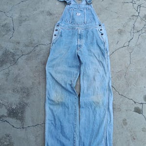 Op de afbeelding: Lichtblauwe denim overall met verstelbare bandjes en meerdere zakken. De overall heeft een klassiek ontwerp met metalen hardware en een kleine patch op de slab. De denim lijkt versleten.