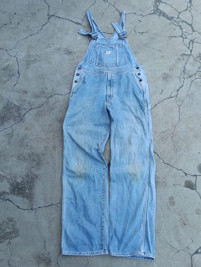 Op de afbeelding: Lichtblauwe denim overall met verstelbare bandjes en meerdere zakken. De overall heeft een klassiek ontwerp met metalen hardware en een kleine patch op de slab. De denim lijkt versleten.
