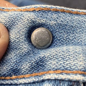 Puede incluir: Primer plano de un bot&oacute;n de metal plateado en unos vaqueros azules. El bot&oacute;n tiene texto en relieve y est&aacute; rodeado de costuras naranjas. La tela vaquera tiene un aspecto texturizado.