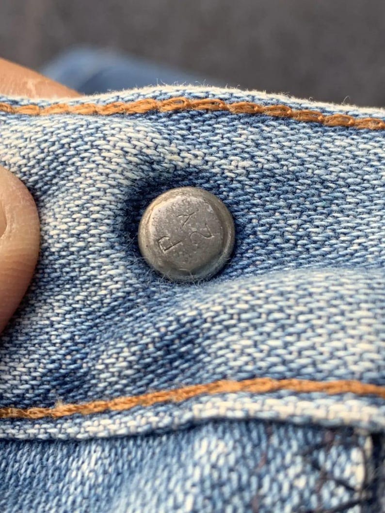 Puede incluir: Primer plano de un bot&oacute;n de metal plateado en unos vaqueros azules. El bot&oacute;n tiene texto en relieve y est&aacute; rodeado de costuras naranjas. La tela vaquera tiene un aspecto texturizado.