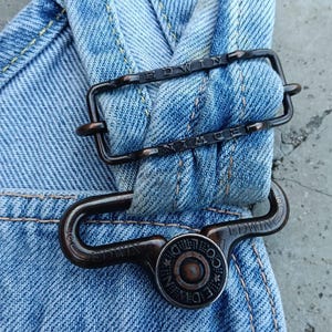 Op de afbeelding: Close-up van een lichtblauwe denim overallband met een donkere metalen gesp en knoop. De gesp en knoop hebben de merknaam "EDWIN" gegraveerd. De denim heeft gele stiksels.