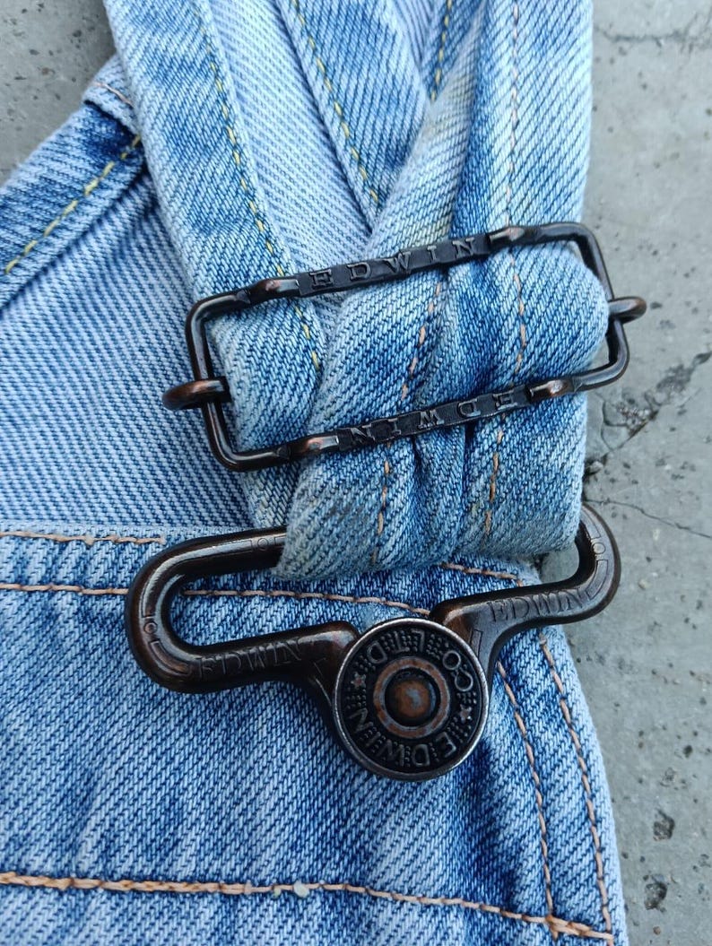 Op de afbeelding: Close-up van een lichtblauwe denim overallband met een donkere metalen gesp en knoop. De gesp en knoop hebben de merknaam "EDWIN" gegraveerd. De denim heeft gele stiksels.