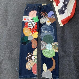 May include: Dark blue denim jeans with colorful patchwork flower designs on the legs. The patches include various patterns and fabrics. An American flag is draped in the background. A unique and artistic garment.