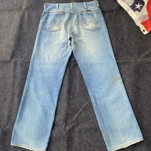 Puede incluir: Vaqueros de mezclilla azul claro con un dise&ntilde;o cl&aacute;sico de cinco bolsillos. Los vaqueros tienen un lavado descolorido y una pernera recta. Una bandera americana es visible en el fondo. Los vaqueros son de estilo vintage.