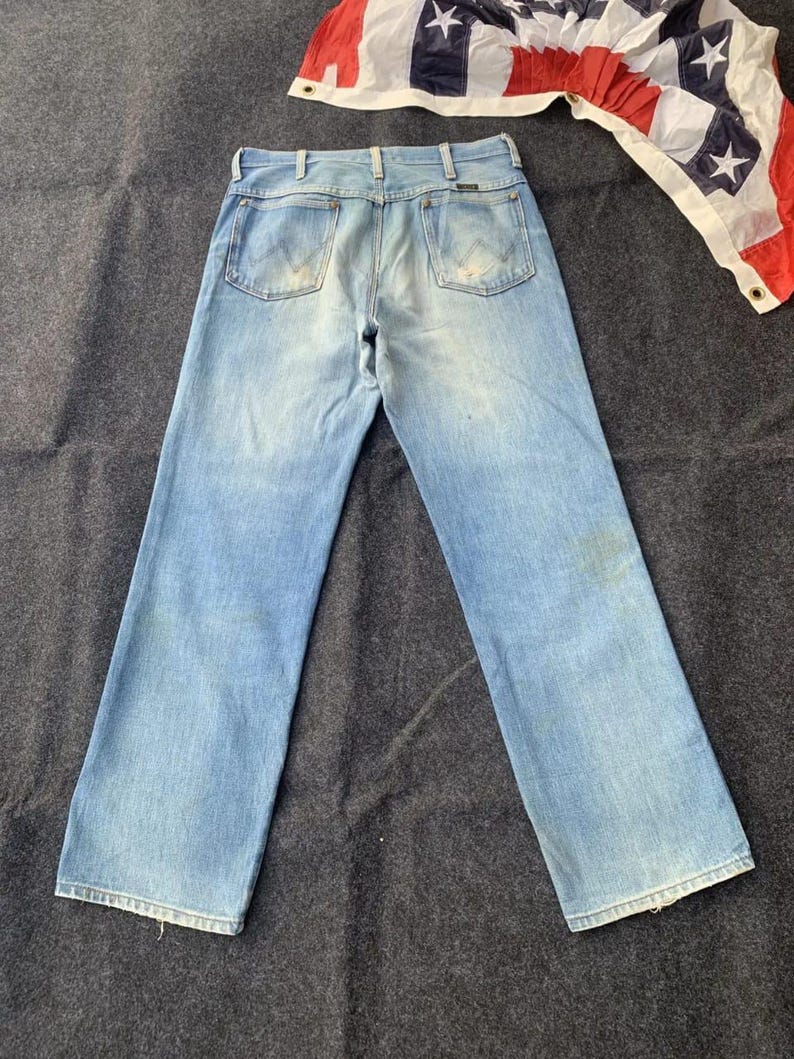 Puede incluir: Vaqueros de mezclilla azul claro con un dise&ntilde;o cl&aacute;sico de cinco bolsillos. Los vaqueros tienen un lavado descolorido y una pernera recta. Una bandera americana es visible en el fondo. Los vaqueros son de estilo vintage.