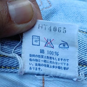 Op de afbeelding: Close-up van een wit label met de tekst "HT4065" en Japanse karakters. Het label bevat ook wasvoorschriften en de tekst "綿 100%", wat 100% katoen aangeeft. Het label is bevestigd aan lichtblauwe denim.