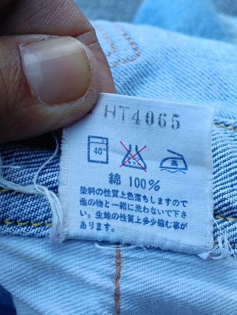 Op de afbeelding: Close-up van een wit label met de tekst "HT4065" en Japanse karakters. Het label bevat ook wasvoorschriften en de tekst "綿 100%", wat 100% katoen aangeeft. Het label is bevestigd aan lichtblauwe denim.