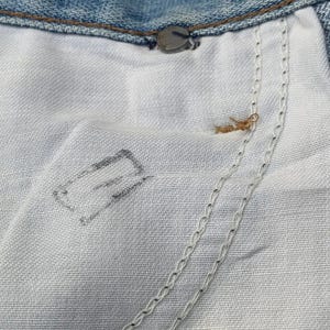 Puede incluir: Primer plano del interior de unos vaqueros azules. La imagen muestra la tela clara del forro del bolsillo, las costuras y el bot&oacute;n y remache met&aacute;licos.