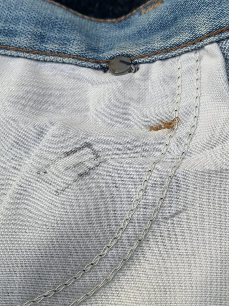 Puede incluir: Primer plano del interior de unos vaqueros azules. La imagen muestra la tela clara del forro del bolsillo, las costuras y el bot&oacute;n y remache met&aacute;licos.