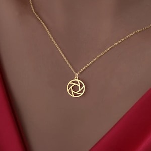 May include: A gold chain necklace with a circular pendant featuring a geometric design resembling a camera aperture.