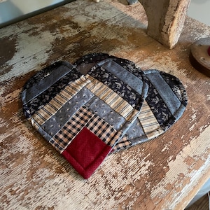 Puede incluir: Tres salvamanteles acolchados en forma de corazón. Los salvamanteles presentan un diseño de patchwork con varias telas en tonos azules, negros y crema. Un cuadrado de tela roja se encuentra en la parte inferior de cada corazón. Los salvamanteles están sobre una superficie de madera envejecida.