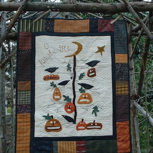 Op de afbeelding: Een handgemaakte quilt met Halloween-thema, met een ontwerp van een pompoenboom met kraaien en pompoen lantaarns. De quilt heeft een crèmekleurige achtergrond, een zwarte rand en patchwork randen. De tekst "O pumpkin tree" staat op de quilt.