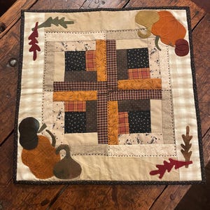 May include: A square fabric wall hanging featuring a central log cabin quilt block pattern in shades of black, brown, and orange. The border is cream-coloured plaid with appliquéd felt pumpkins and autumn leaves in the corners, finished with dark trim.