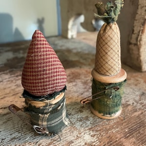 May include: Two handmade fabric gnome ornaments. One has a red and white checkered hat, the other has a tan and brown plaid hat. Both have green and tan plaid skirts and are attached to wooden spools with safety pins. Rustic home decor.