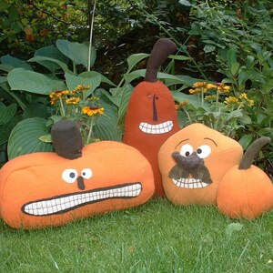 May include: A collection of four orange fabric pumpkin-shaped plush toys with brown stems. Each plush features a unique embroidered face with white teeth, black eyes, and one figure includes a brown fabric moustache and nose detail.