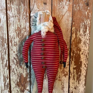 Peut inclure: Une poupée en tissu représentant le Père Noël, vêtue d'une combinaison rayée rouge et beige avec de petites clochettes en métal sur le devant. La poupée porte un long bonnet de nuit assorti, une barbe blanche duveteuse, des mains et des pieds noirs.