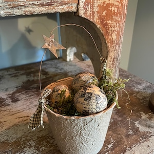 May include: A rustic, textured paper pot filled with moss and three spherical ornaments covered in vintage newspaper print. A rusty metal star on a wire extends above the pot, and a small plaid bow adds a decorative touch. The arrangement sits on a distressed wooden surface.