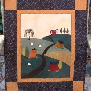 May include: A handcrafted quilt featuring a rural landscape scene. The quilt showcases a farm with rolling hills, a winding road, and several buildings in shades of red, orange, and brown. The border is a dark blue with small tan dots and tan corner squares.