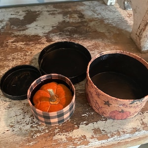 May include: Two round, open-top containers with black interiors. One smaller container features a black and orange buffalo check pattern and holds a small orange fabric pumpkin. The larger container has a tan exterior with a pumpkin and star print.